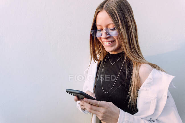 Mujer sonriente con gafas de sol elegantes navegando por el teléfono móvil de pie cerca de la pared blanca y mirando a la pantalla - foto de stock