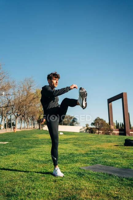 Der erwachsene männliche Athlet in Sportkleidung hebt das Bein und freut sich beim Training auf dem Rasen im Sonnenlicht — Stockfoto