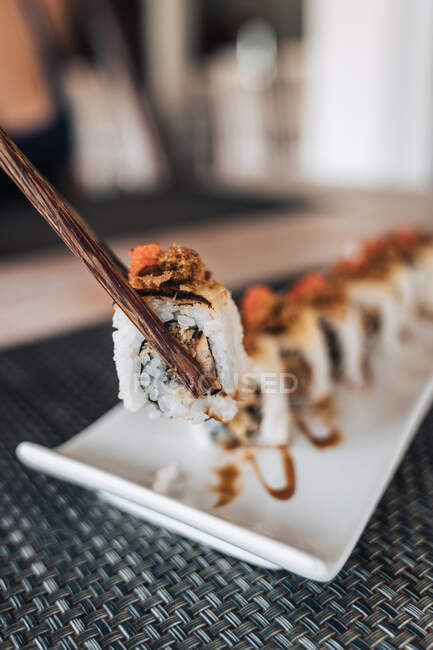 Pessoa de colheita segurando com pauzinhos fileira de saborosos rolos de sushi com arroz cozido e fatias de frutos do mar em prato cerâmico na mesa — Fotografia de Stock