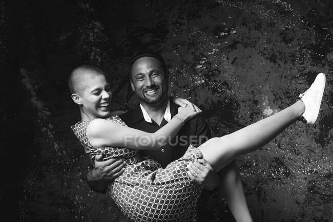 Черно-белый образ жизнерадостного мужчины, держащего подругу и веселящегося в темной студии — стоковое фото