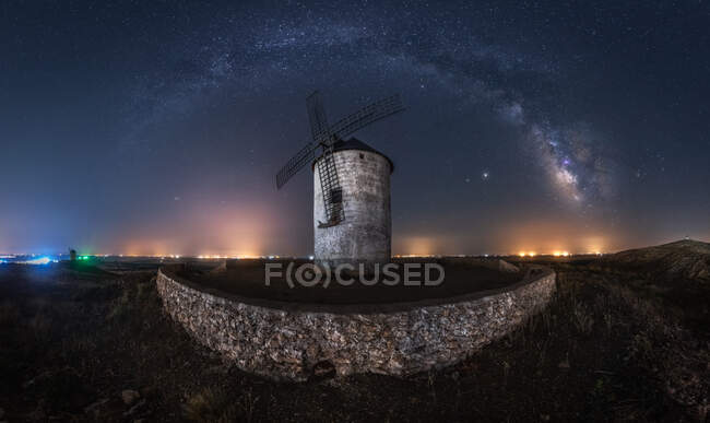 Picturesque scenery of Milky Way in dark night sky above aged stone windmill tower with glowing lights in distance — Stock Photo