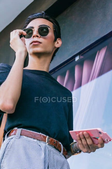 Desde abajo de hombre gay en gafas de sol de moda y con teléfono móvil de pie en la ciudad y mirando hacia otro lado - foto de stock