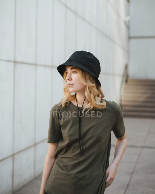 Young female in casual apparel looking away against concrete wall of modern building on urban pavement in daytime — Stock Photo