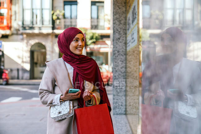 Délicieuse femme musulmane acheteuse en foulard et avec des sacs à provisions debout près de la vitrine du magasin en ville — Photo de stock