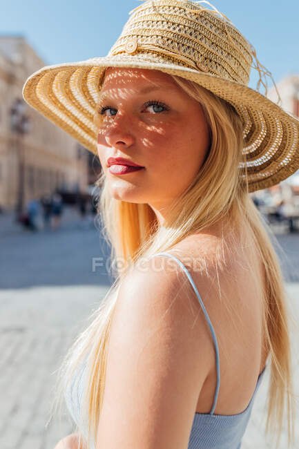 Вид сбоку очаровательной женщины в соломенной шляпе, смотрящей в камеру в солнечный день на городской улице летом — стоковое фото