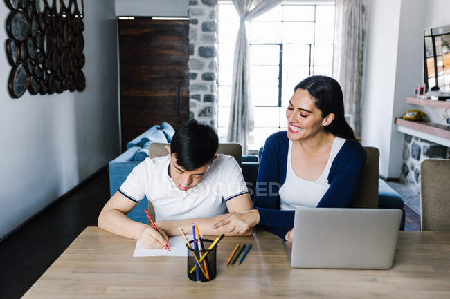 Етнічний хлопчик-підліток з синдромом Дауна малює олівцями на папері, сидячи за столом з фрілансером, який працює на ноутбуці вдома — стокове фото