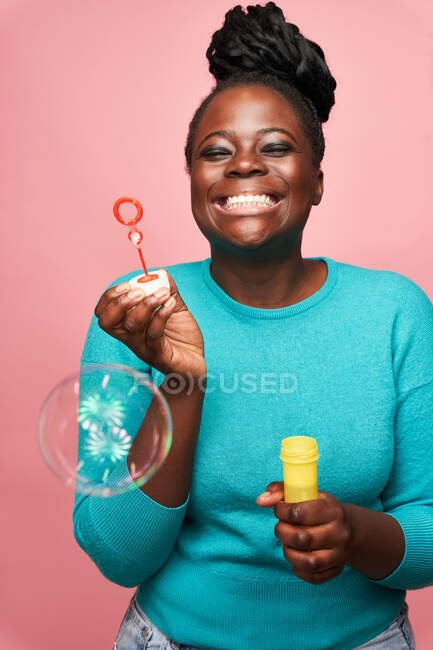 Happy African American female with eyes closed wearing blue clothes and blowing soap bubbles against pink background in studio — Stock Photo