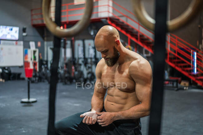 Deportista calvo sin camisa revisando las manos lesionadas cubiertas de tiza mientras está sentado en el gimnasio durante el entrenamiento funcional - foto de stock