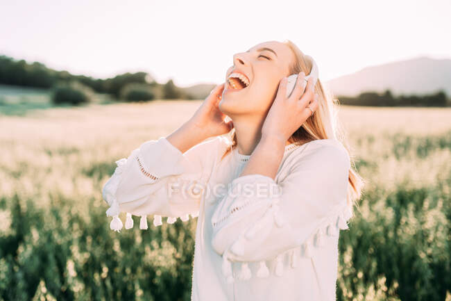 Eccitato femmina in abito bianco con frange e ascoltare musica con cuffie cantare con gli occhi chiusi sul campo di campagna — Foto stock