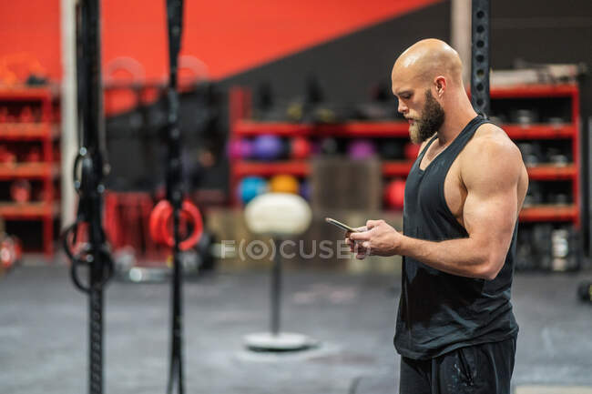 Vue latérale du sportif chauve musclé debout dans la salle de gym moderne naviguant sur smartphone pendant la pause d'entraînement — Photo de stock