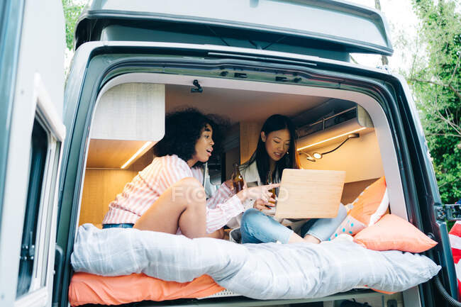 Jóvenes amigas multirraciales bebiendo cerveza y usando el portátil juntas mientras preparan contenido para el blog de viajes dentro de la caravana durante la aventura de verano en la naturaleza - foto de stock