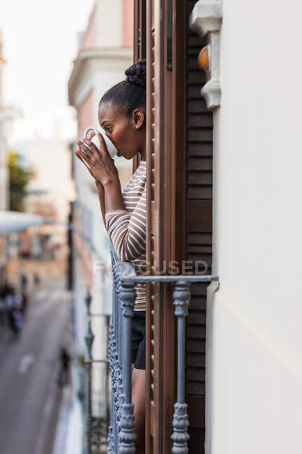 Vista laterale della donna afroamericana riflettente con tazza di bevanda calda guardando lontano dal balcone in città — Foto stock