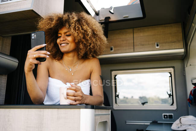 Felice donna afroamericana con tazza di bevanda calda sorridente e navigando cellulare mentre riposava nella carovana contemporanea al mattino — Foto stock