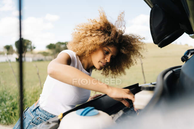Side view of traveling female looking under opened hood of camper van while having problems during trip through summer nature — Stock Photo