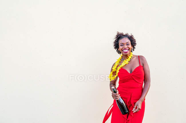 Популярна афроамериканська жінка в квітковому намисті, одягнена в яскраву загальну форму, стоїть з пляшкою шампанського на білому тлі. — стокове фото