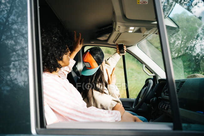 Through window side view of cheerful young female friends having fun and taking selfie on smartphone while sitting inside camper vehicle during summer trip in nature — Stock Photo