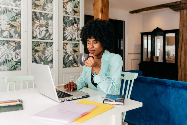 Jeune pigiste afro-américaine en tenue décontractée buvant du café assis à table et travaillant sur un projet avec ordinateur portable à la maison — Photo de stock