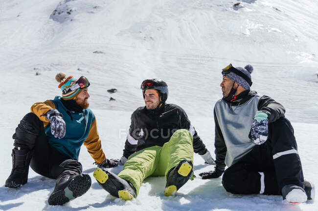 Улыбающиеся бородатые спортсмены в спортивной одежде разговаривают сидя на снегу и глядя друг на друга против Сьерра-Невады — стоковое фото