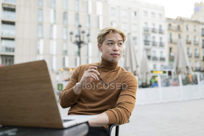 Pensive Asian male freelancer sitting with laptop at table in street cafe and working remotely on startup while looking away — Stock Photo