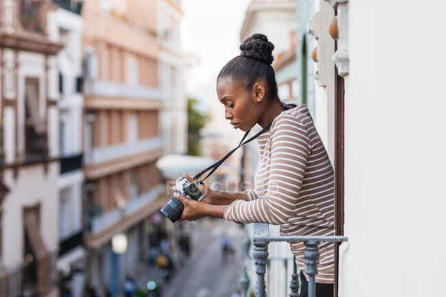 Side view of young African American woman with professional photo camera standing on fenced balcony in town — Stock Photo