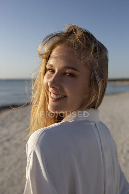 Mujer rubia con el pelo largo de pie en la playa mirando la cámara - foto de stock