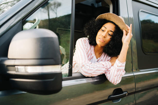 Joven mujer afroamericana positiva en traje de verano y sombrero inclinado por la ventana de la caravana y mirando al espejo lateral mientras disfruta del viaje por carretera - foto de stock