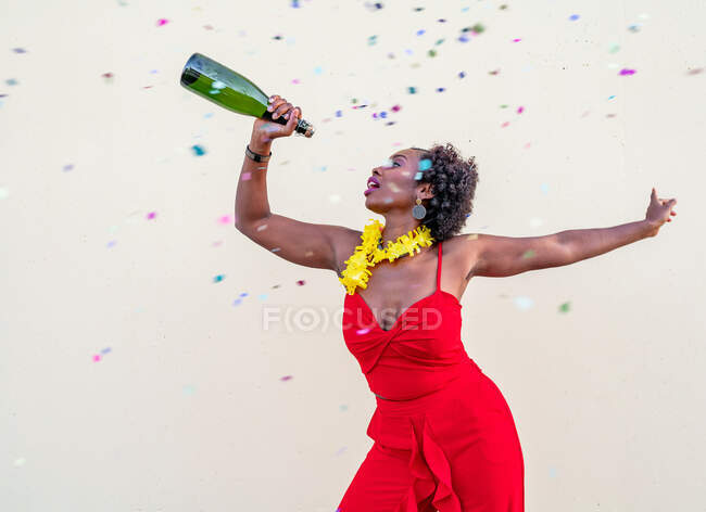 Популярна афроамериканська жінка в квітковому намисті, одягнена в яскраву загальну форму, стоїть з пляшкою шампанського на білому тлі. — стокове фото