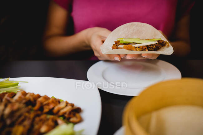 Hands of woman sitting at dinner table and holding Beijing duck piece on rice flat bread over ceramic plate — Stock Photo