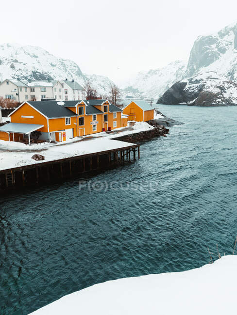 Cottage gialli e banchina innevata situati vicino al mare increspato contro le montagne nella fredda giornata invernale nel villaggio costiero sulle isole Lofoten, Norvegia — Foto stock