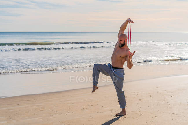 Vista lateral del atleta masculino sin camisa estirando los brazos con banda elástica mientras está de pie en una pierna haciendo ejercicio en la playa soleada vacía - foto de stock