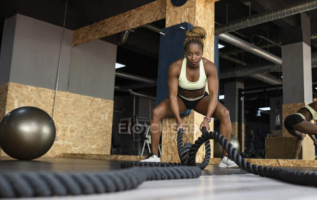 Atleta afro-americana forte em activewear exercitando-se com cordas de batalha enquanto olha para a câmera durante o treinamento de alta intensidade no ginásio — Fotografia de Stock