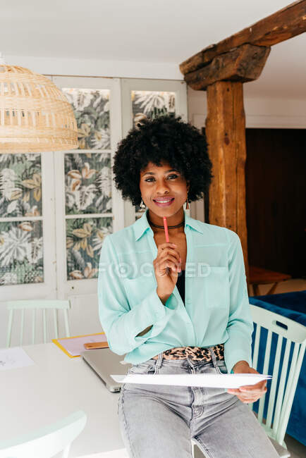 Moderna donna afro-americana freelance di successo in abito elegante con capelli afro sorridente guardando la fotocamera mentre si siede a tavola e leggere il documento a casa — Foto stock