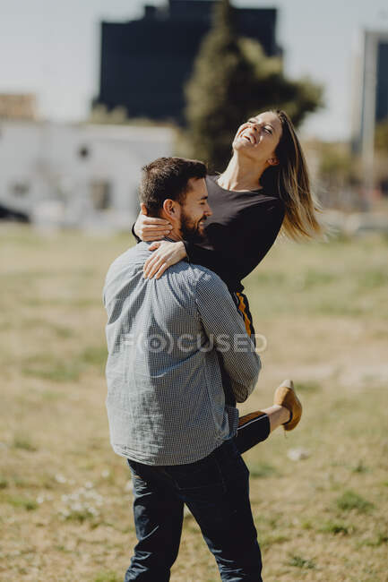 Бородатый мужчина обнимает и поднимает смеющуюся женщину, проводя время на зеленой лужайке на улице вместе — стоковое фото