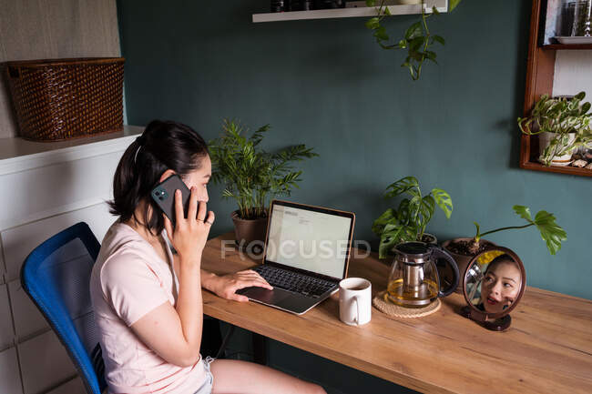 Vista lateral de la mujer freelancer étnica enfocada que tiene conversación en el teléfono móvil mientras navega por la computadora portátil y trabaja en el proyecto en línea desde casa - foto de stock