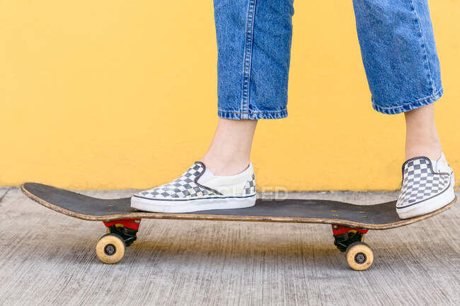 Cropped unrecognizable female skater with skateboard standing on walkway with colorful yellow wall on the background in daytime — Stock Photo