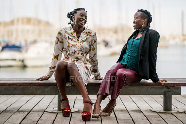 Élégantes dames afro-américaines souriantes à la mode passant du temps ensemble assis sur un banc bas en bois dans le parc par temps clair — Photo de stock