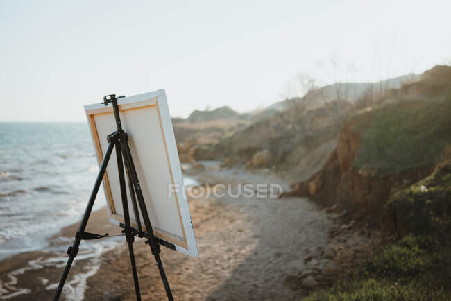 Painting on easel placed on sandy beach washing by foamy sea surrounded by rocky cliffs on sunny day — Stock Photo