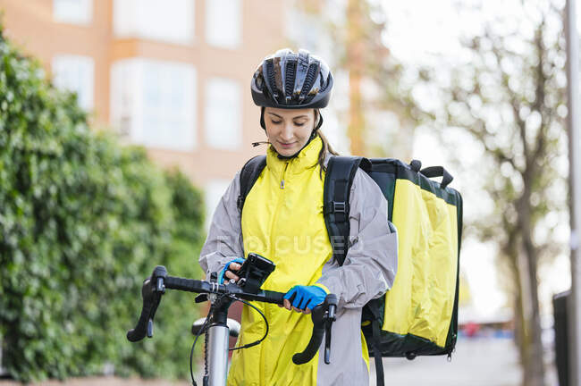 Young woman with thermal bag browsing smartphone with GPS map on bicycle handlebar while delivering food on city street — Stock Photo