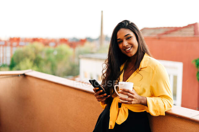 Ottimista donna ispanica con tazza di bevanda calda sorridente e guardando la fotocamera mentre si appoggia sul balcone ringhiera e navigando cellulare — Foto stock