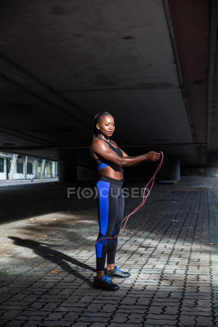 Vista lateral de la hermosa mujer afroamericana en ropa deportiva sosteniendo la cuerda de salto y mirando a la cámara mientras está parado en el pavimento en la calle de la ciudad - foto de stock