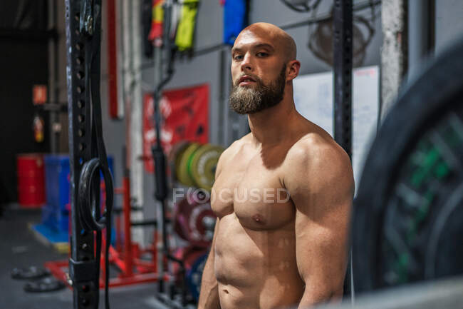 Мускулистый бородатый мужчина смотрит в камеру, стоя рядом с оборудованием во время тренировки в современном тренажерном зале — стоковое фото