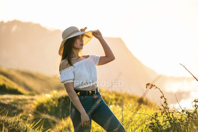 Attractive dreamy female in jeans and white top touching hat brim and looking away in pleasant thoughts while standing on lush grassy terrain on sunny day — Stock Photo