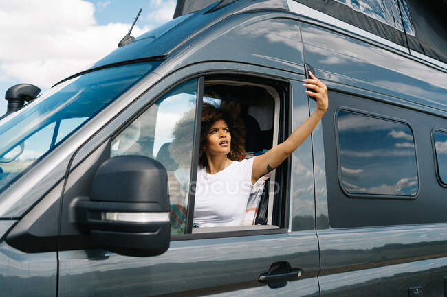 Unhappy young African American female traveler sitting on driver seat in camper van and trying to get connection on mobile phone while having troubles during road trip — Stock Photo