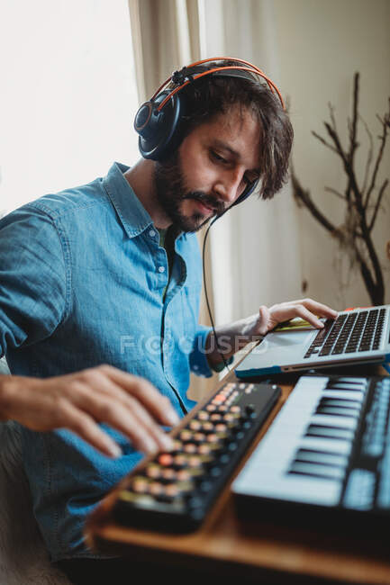 Вид сбоку молодого человека в наушниках с помощью синтезатора и ноутбука за домашним столом — стоковое фото