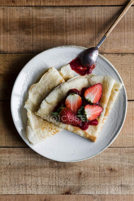 Top view of delicious crepes with sweet strawberry jam placed on plate near spoon on wooden table — Stock Photo