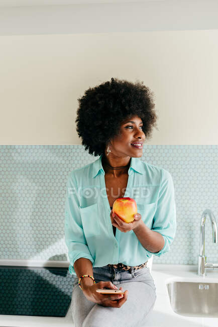 Moderna mujer afroamericana hermosa con teléfono inteligente en la mano sentado en el mostrador de la cocina mirando hacia fuera en casa y comiendo manzana - foto de stock