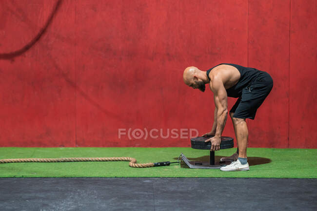 Вид збоку лисий хлопець у спортивному одязі, що кладе важкі пластини на металеву платформу з мотузкою під час функціональних тренувань у спортзалі — стокове фото