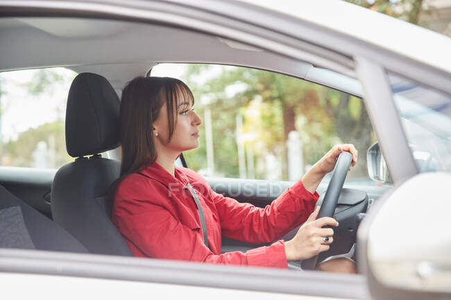 Through window side view of confident young female in red jacket driving modern automobile in city — Stock Photo