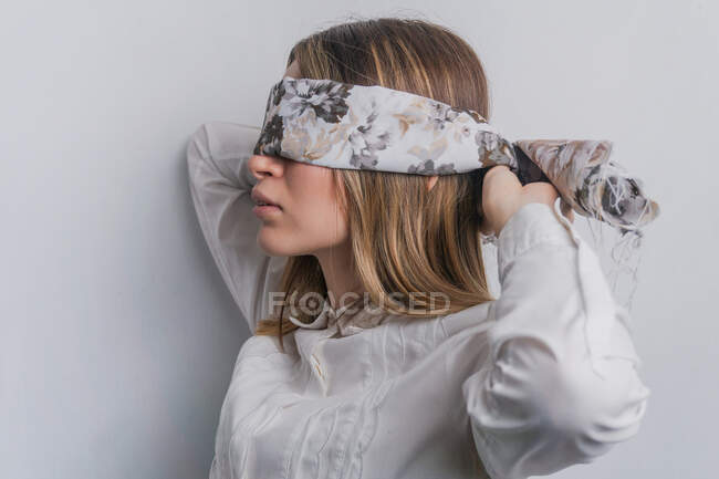 Side view of young fair haired female in elegant blouse covering eyes with folded silk scarf with floral ornament against white background — Stock Photo