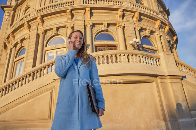 Счастливая взрослая женщина в синем пальто, стоящая рядом со старым зданием, разговаривая по телефону в районе города в солнечный день под голубым облачным небом — стоковое фото
